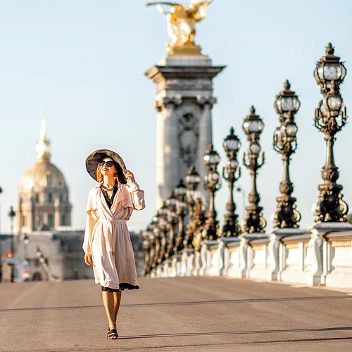 Högtidlig kvinna i elegant klänning och hatt som promenerar på Pont Alexandre III-bron i Paris med utsikt över stadens historiska byggnader och Eiffeltornet i bakgrunden.