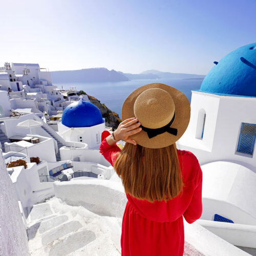 en tjej i knallröd klänning och stråhatt och kikar utöver blåa tak på Santorini
