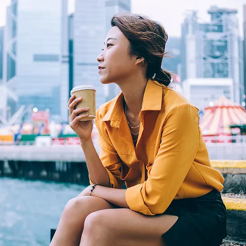 En kvinna njuter av en kopp kaffe med Hong Kongs stadssilhuett i bakgrunden, inklusive höga skyskrapor och hamn. Perfekt för att visa stadens moderna arkitektur och livliga atmosfär.