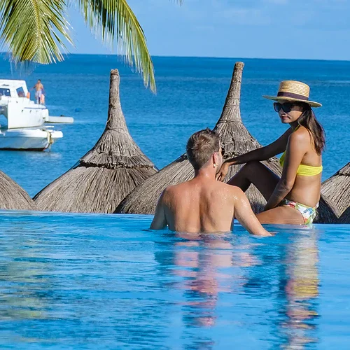 Två personer som njuter av en pool vid stranden på Mauritius, med palmer och båtar i bakgrunden, perfekt för avkoppling och solsemester.