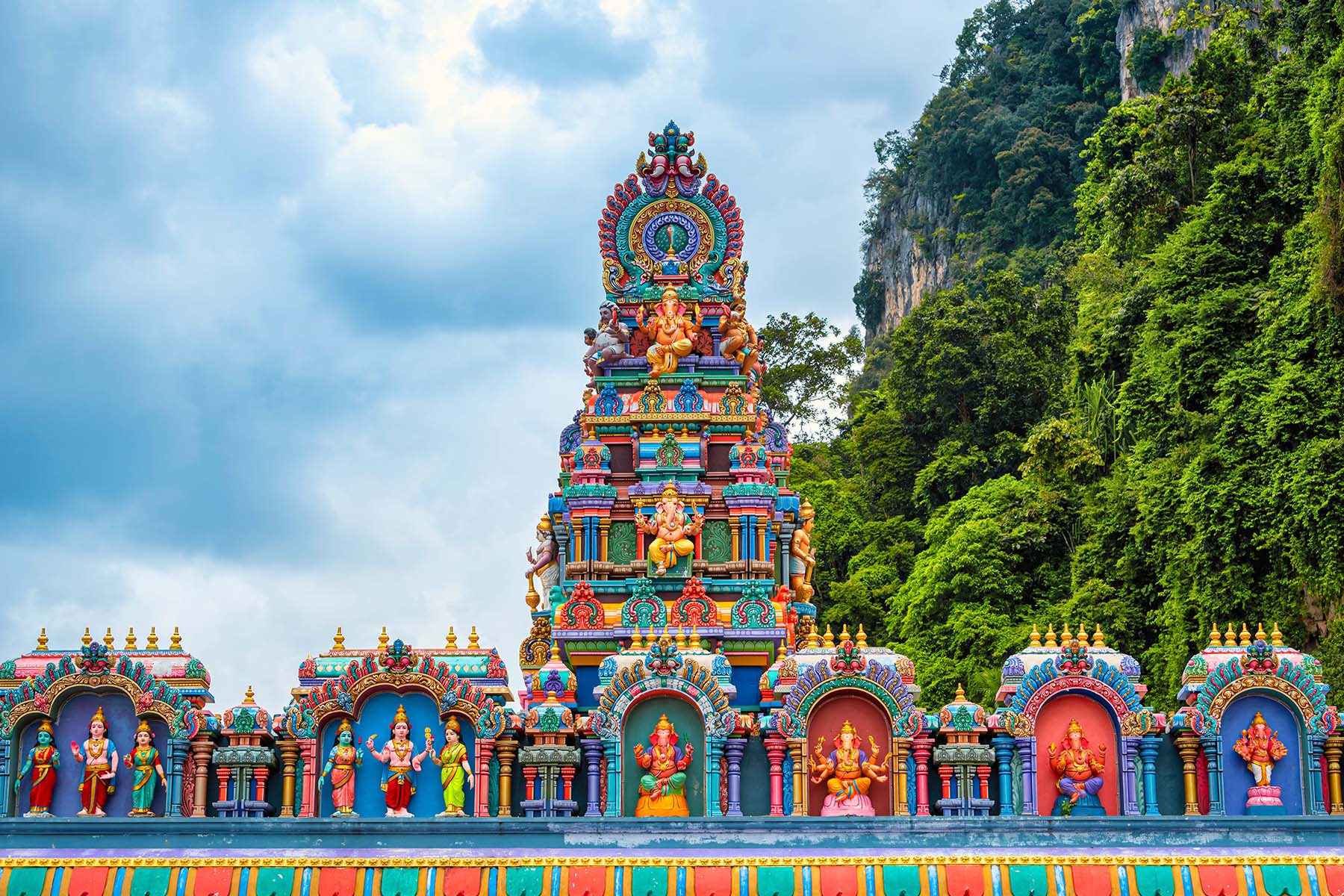 Färgglad hinduiska tempel med höga spiraler och detaljerade statyer av gudar, omgivet av gröna berg och molnig himmel i bakgrunden, symboliserar spiritualitet och kultur i Sydostasien.