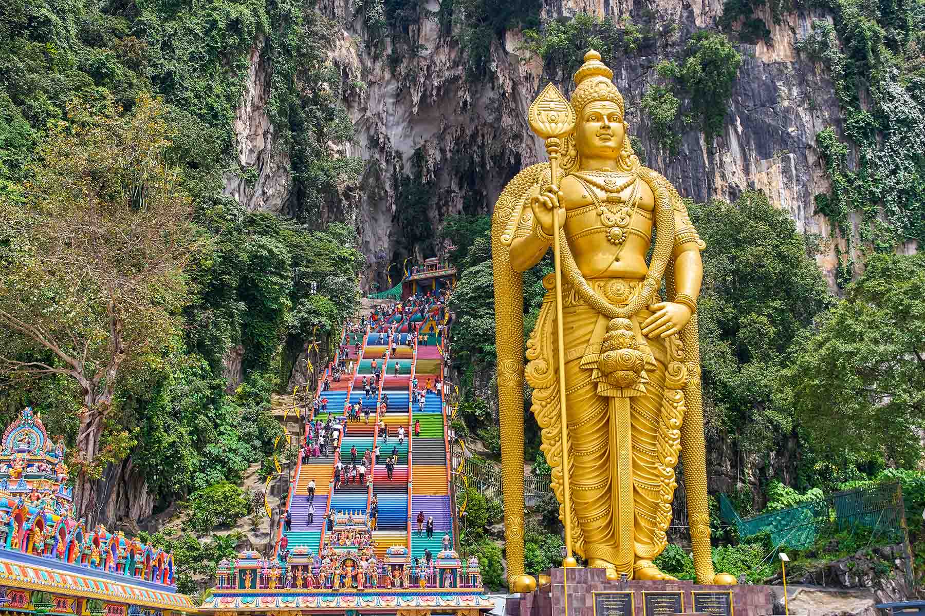 Gyllenstaty av Lord Murugan vid Batu Caves i Malaysia, med färgglada trappor och frodiga gröna omgivningar, symboliserar hinduisk tro och kultur i en historisk helgedom.