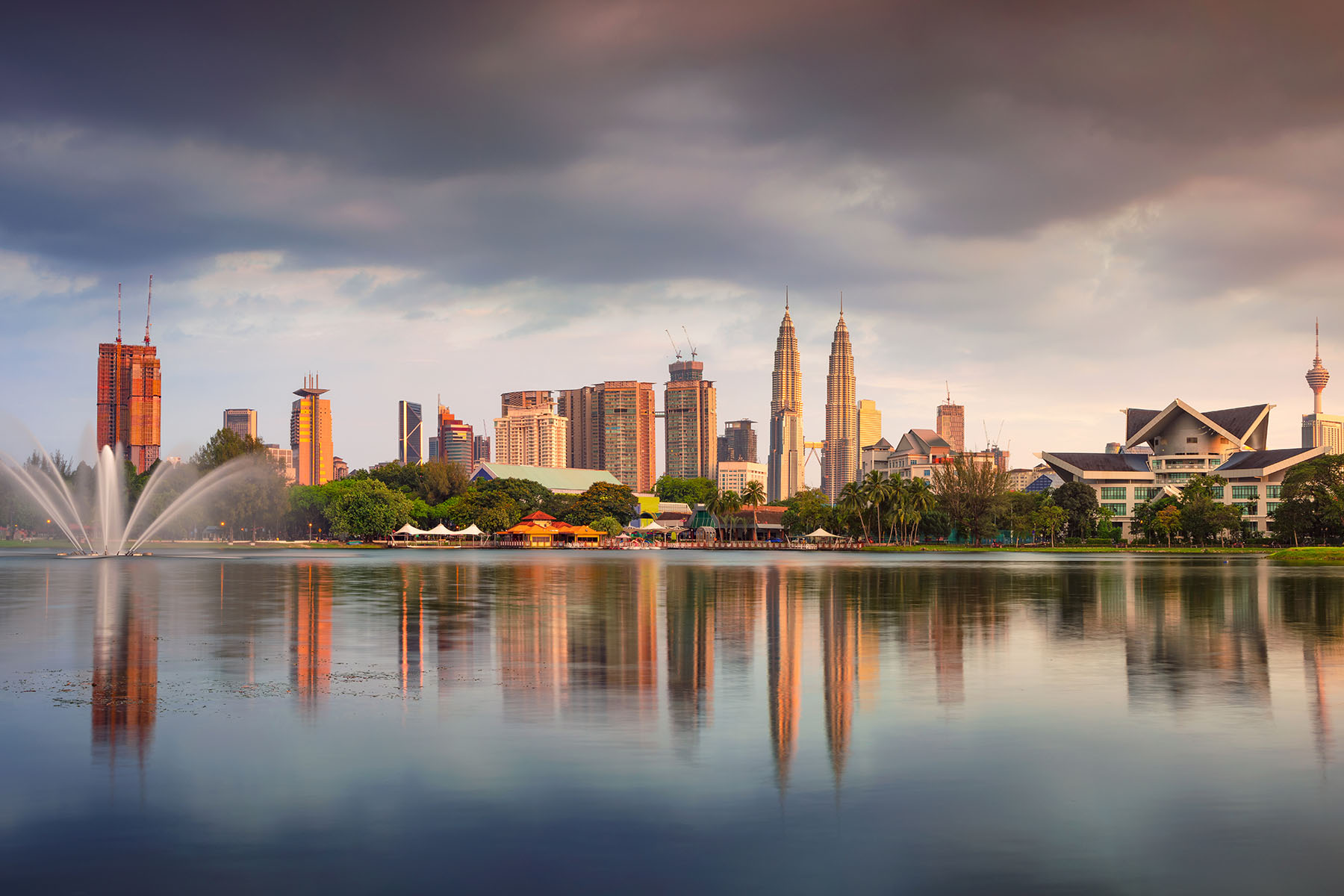 1. Modern Kuala Lumpur skyline med Petronas Twin Towers vid vattnet och en lugn spegling på dammen, under en skyfull molnhimmel i skymningen.