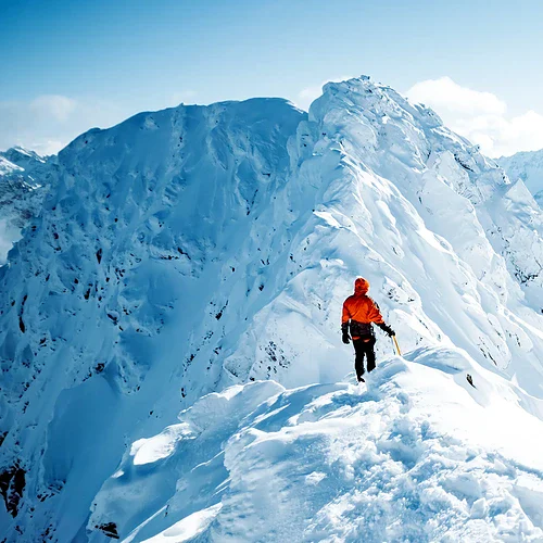 En person i orange jacka vandrar på en snötäckt bergstopp med majestätiska snöklädda berg i bakgrunden, under en klar dag.