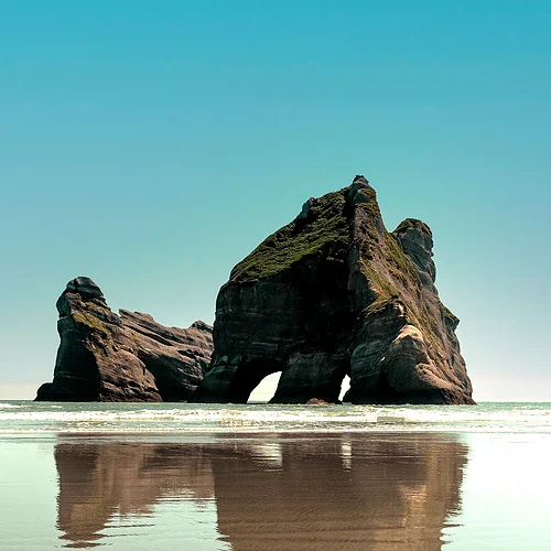 Strand med stor naturlig stenformation och en person som går längs vattnet i Nya Zeeland.