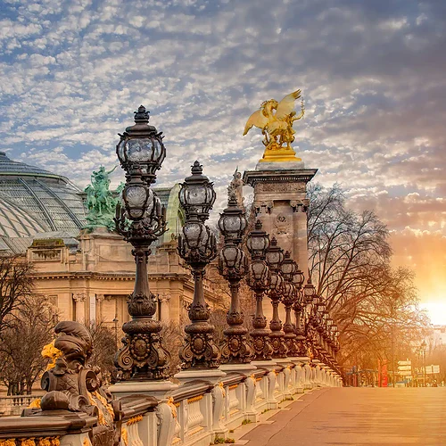 Elegant Parisian boulevard with ornate street lamps, historic statues, and the glass-domed Grand Palais in the background during sunset.
