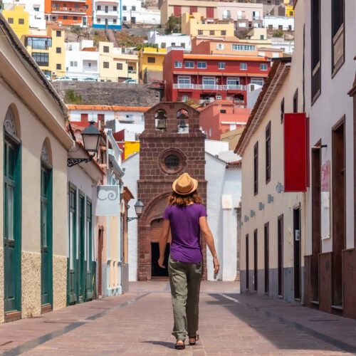 En kvinna med hatt promenerar längs en gata i San Sebastian, omgiven av färgglada byggnader och kullerstensgator, med stadens berg och hus i bakgrunden.
